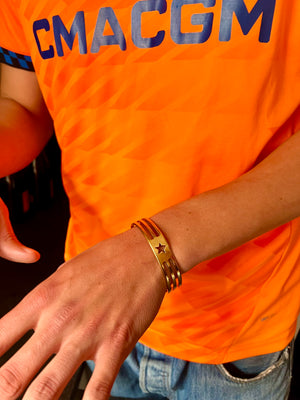 A person in a bright orange CMACGM shirt and blue jeans displays their left wrist adorned with the UNISTAR BRACELET—a handmade gold piece with a star and three lines—while on the streets of Munich.