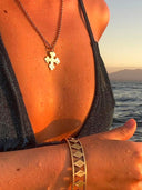 Wearing the Kathl-Gross ORTHO CROSS NECKLACE, a person in a wet dark swimsuit and geometric bracelet stands near water, sunlight highlighting droplets on their skin with mountains visible in the background.