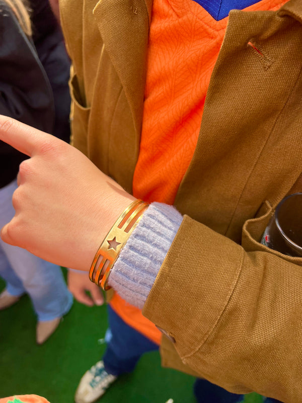 In Munich, someone in a brown jacket, orange sweater, and blue shirt gestures outdoors with a black cup. They wear the handmade UNISTAR BRACELET—a gold piece adorned with charming star cut-outs and stripe designs.