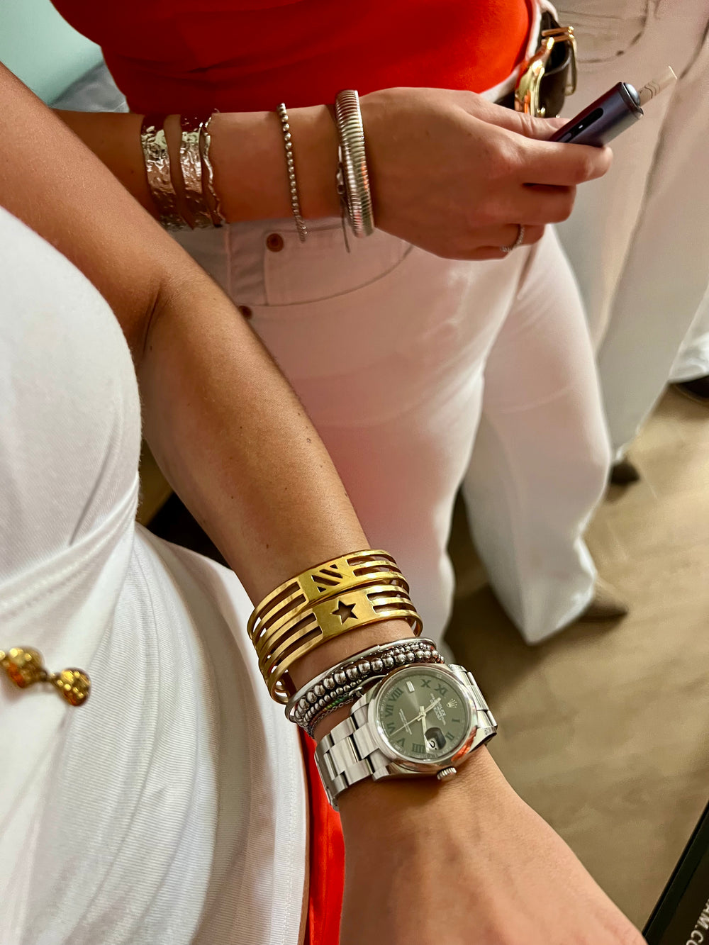 A person in a bright orange CMACGM shirt and blue jeans displays their left wrist adorned with the UNISTAR BRACELET—a handmade gold piece with a star and three lines—while on the streets of Munich.