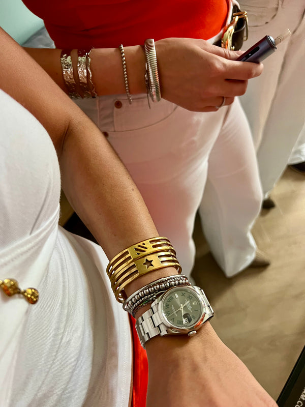 Two people in white outfits, seen from the shoulders down. One holds a phone and wears silver bangles; the other sports handmade jewelry, including a gold cuff, silver watch, gold beaded bracelet, and the UNISTAR BRACELET. Spotted in Munich.