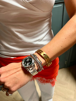 In Munich, a person in a white top and red pants with lace trim showcases handmade jewelry, including the UNISTAR BRACELET, a silver wristwatch, multiple silver bracelets, and a gold cuff with a star cut-out.