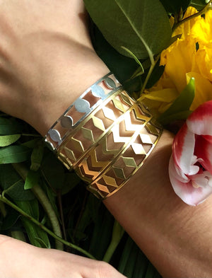 A close-up of a wrist wearing the PATTERN BRACELET SET—four gold geometric bangles and a silver disc bracelet with circular cutouts—surrounded by green leaves and vibrant flowers.