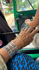 A close-up of a hand wearing the PATTERN BRACELET SET and a patterned ring rests inside a green auto rickshaw, while someone in a sleeveless top drives with greenery and the road visible outside.