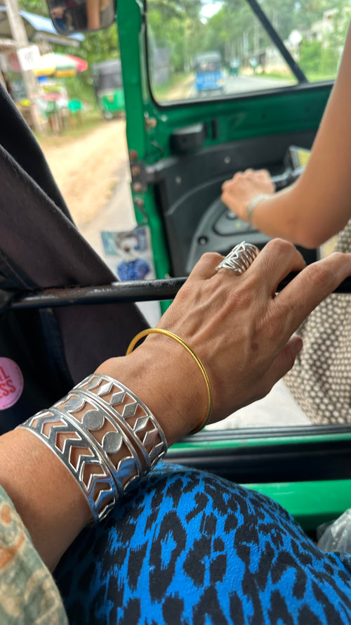 A close-up of a hand wearing the PATTERN BRACELET SET and a patterned ring rests inside a green auto rickshaw, while someone in a sleeveless top drives with greenery and the road visible outside.