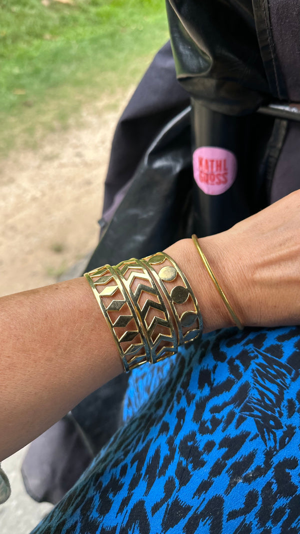 A person wears the PATTERN BRACELET SET, featuring three gold geometric bangles, a thin gold bracelet, and an arrow bracelet, with blue and black fabric below and a stroller handle labeled KATHI CROSS in the background.