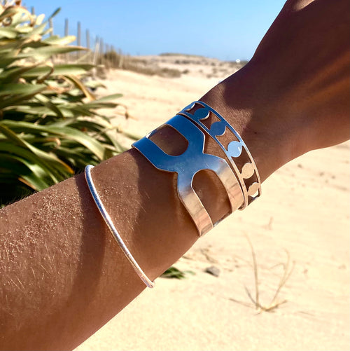 A tanned arm wears three DISC BRACELETS—each 1.5 cm wide with organic silver patterns—set against sand, greenery, and blue sky for handmade Munich charm at the beach.