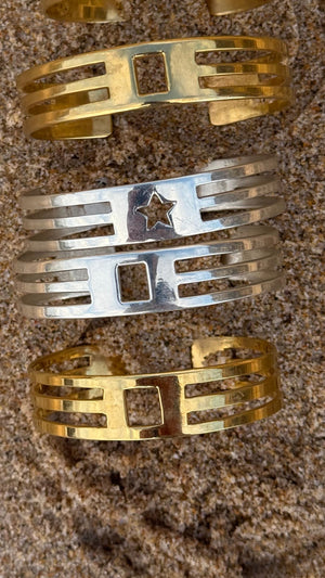 The UNISTAR BRACELET collection features three handmade metallic cuffs—two gold and one silver with a central star cut-out—displayed on coarse sand and inspired by Munich style, each showcasing modern geometric designs.