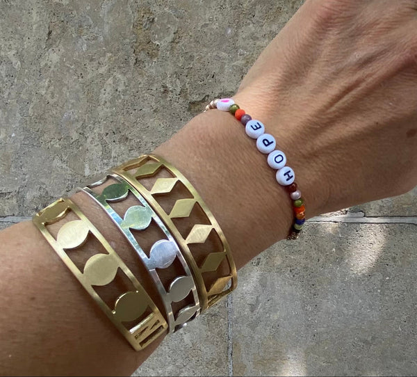 A hand wears three metallic DISC BRACELETs in gold and silver, plus a colorful beaded bracelet with white letter beads spelling HOPE, 1.5 cm wide, handmade in Munich—all set against a stone background.