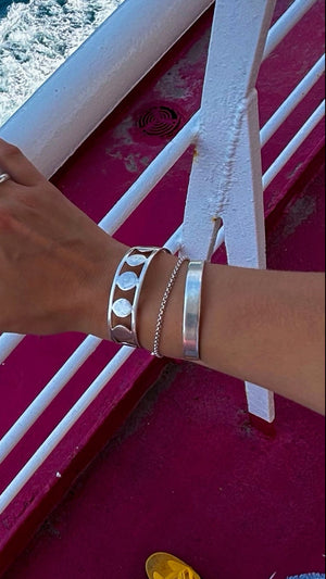 A wrist adorned with three silver handmade DISC BRACELETS, each 1.5 cm wide and featuring organic patterns, is shown against a bright pink background and white railing; the ocean and yellow shoes are also partially visible.