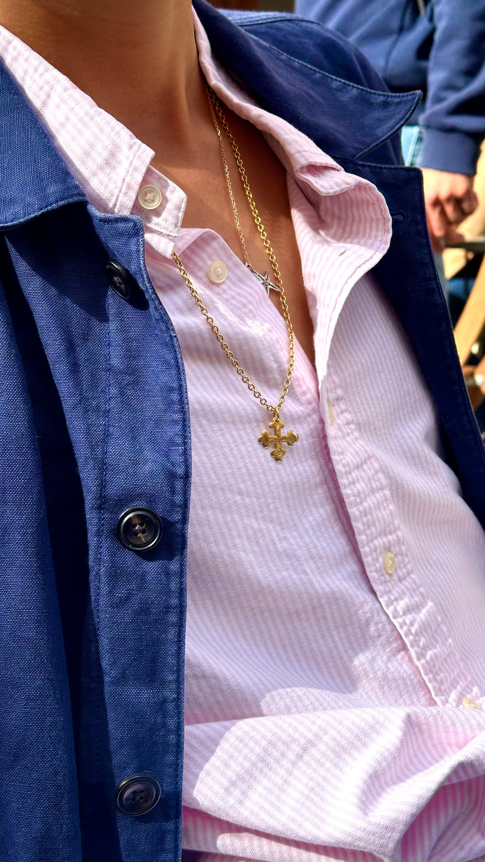 Wearing the Kathl-Gross ORTHO CROSS NECKLACE, a person in a wet dark swimsuit and geometric bracelet stands near water, sunlight highlighting droplets on their skin with mountains visible in the background.