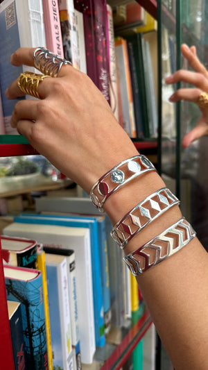 A hand reaches for books on a shelf, wearing the PATTERN BRACELET SET—two silver geometric bracelets—with gold rings and an arrow bracelet. Colorful books fill the background.