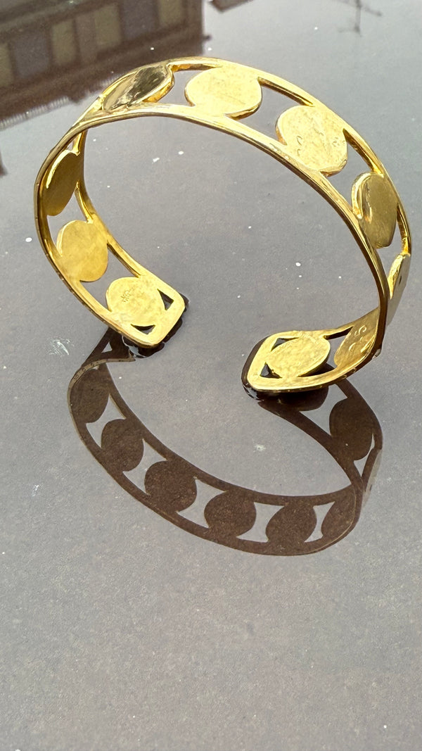 The DISC BRACELET, a 1.5 cm wide gold cuff with circular disc designs, stands upright on a reflective surface, its shadow forming an organic pattern that mirrors the bracelet’s shape.