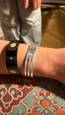 In Munich, a close-up shows a wrist wearing the handmade UNISTAR BRACELET—a silver piece with a star cut-out and three parallel bars—beside a black floral bracelet, set against patterned fabric.