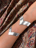 The LOVE CUFF by Kathl-Gross—a silver, angular bracelet handmade in Munich with a striking 10 cm width—wraps elegantly around the forearm, set against a background of patterned, ornate red and gold fabric.